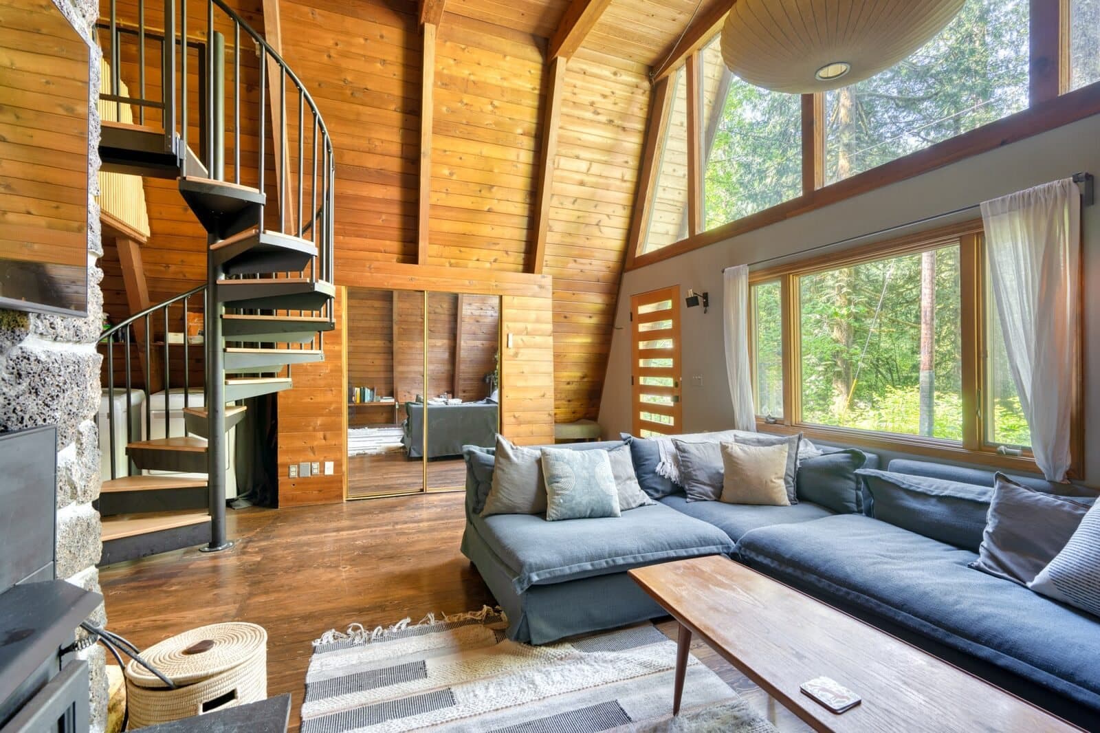Listing photo: inside the A-frame, a living room with a spiral stair and tall windows onto the forest